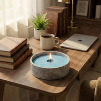 Candle in a decorative bowl on a wooden table with books, a laptop, and a plant.